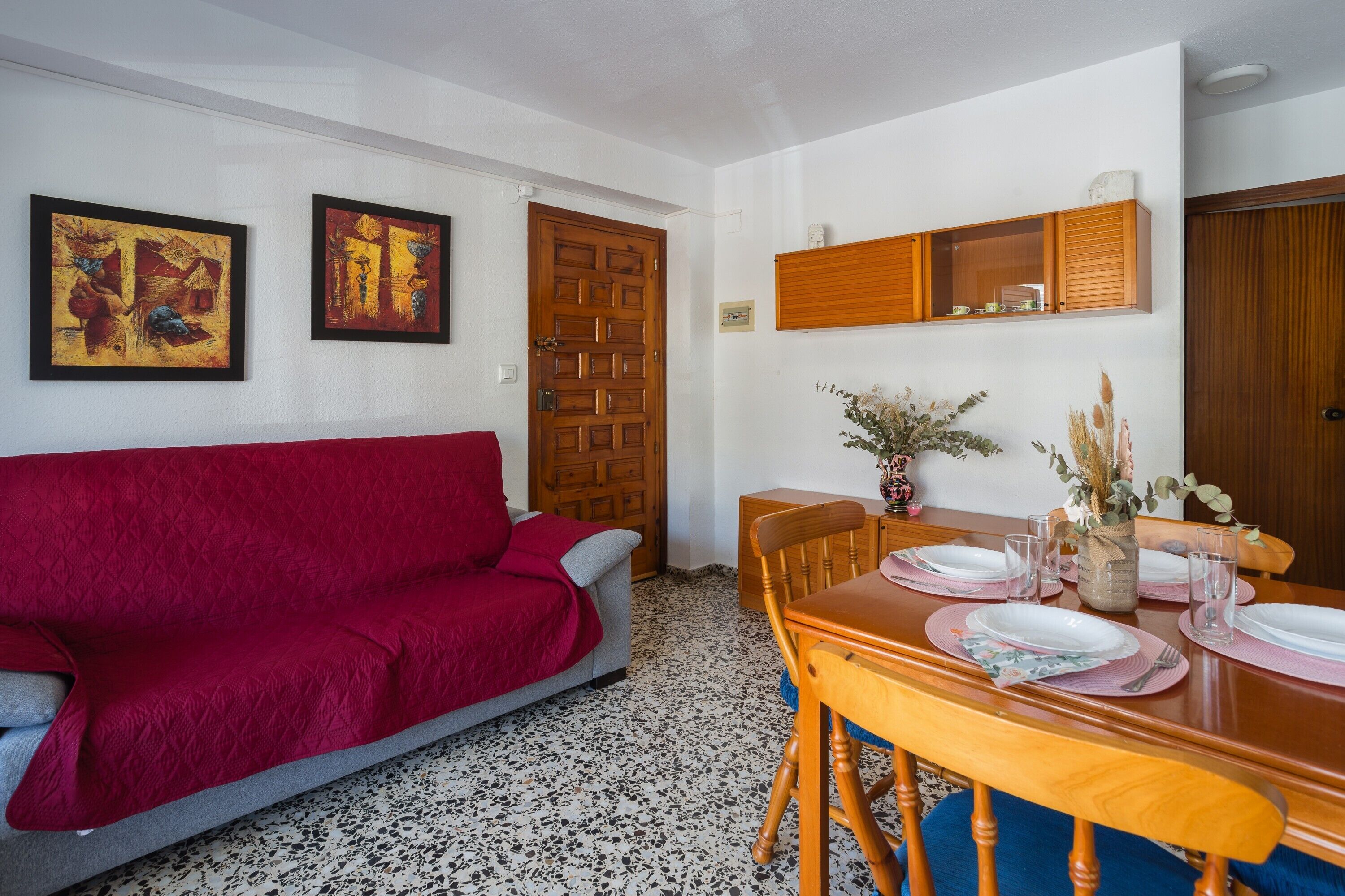 A dining area with a wooden table set for four, a red sofa, and two framed artworks on the wall.