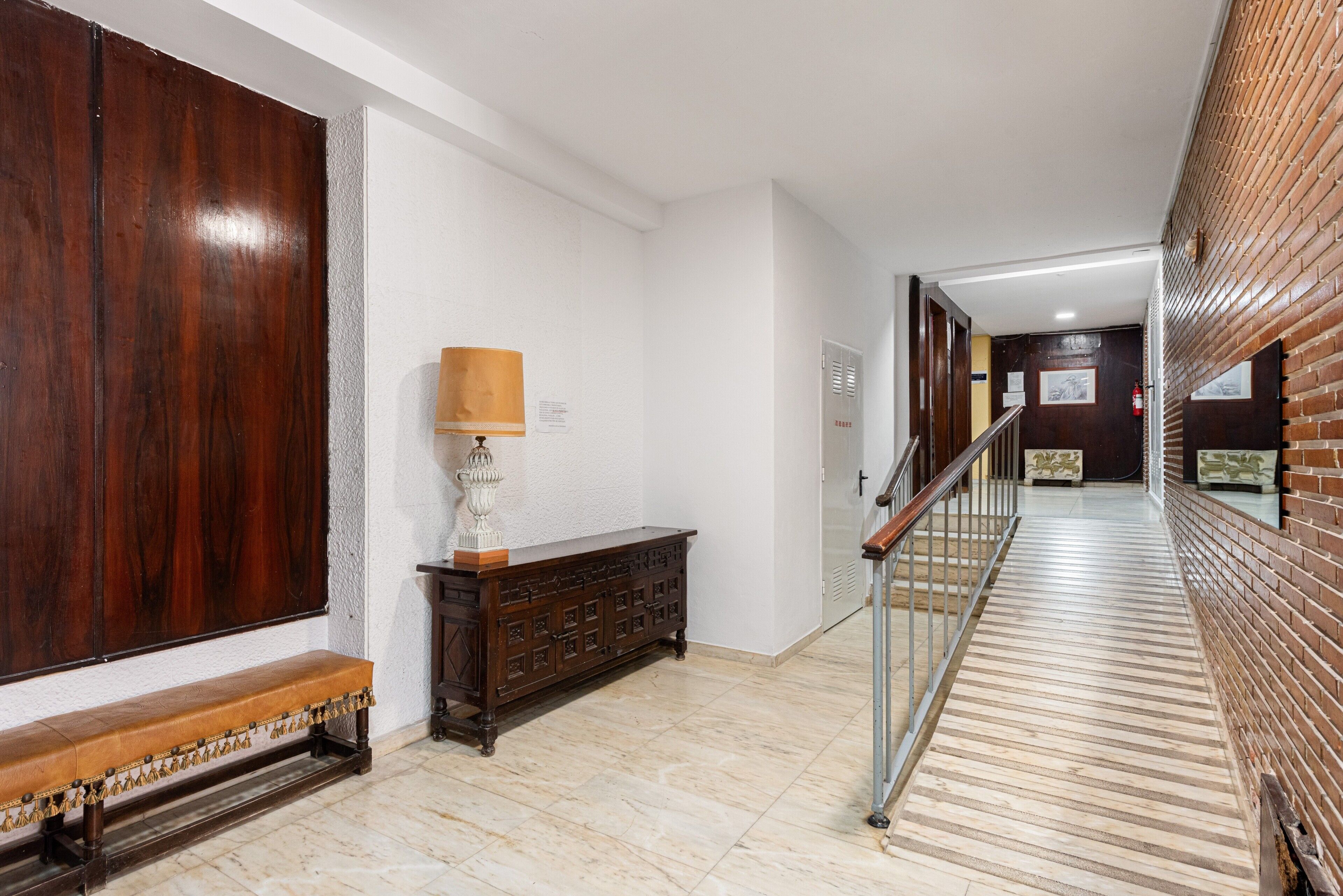 Um corredor com um banco de madeira, uma luminária, um aparador de madeira e uma escada com corrimão de metal.
