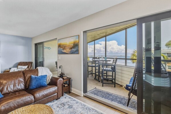 A living room with a brown leather sofa, a glass door leading to a balcony with a view of the sea.