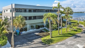 A two-story building with a parking area, palm trees, and a view of the water. - Bayview Condo, Unit E - Anna Maria Island - 2 Bedroom/ 2 Bath bay view, maximum occupancy of 4 people. (Bradenton Beach)