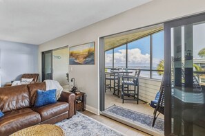A living room with a brown leather sofa, a glass door leading to a balcony with a view of the sea.