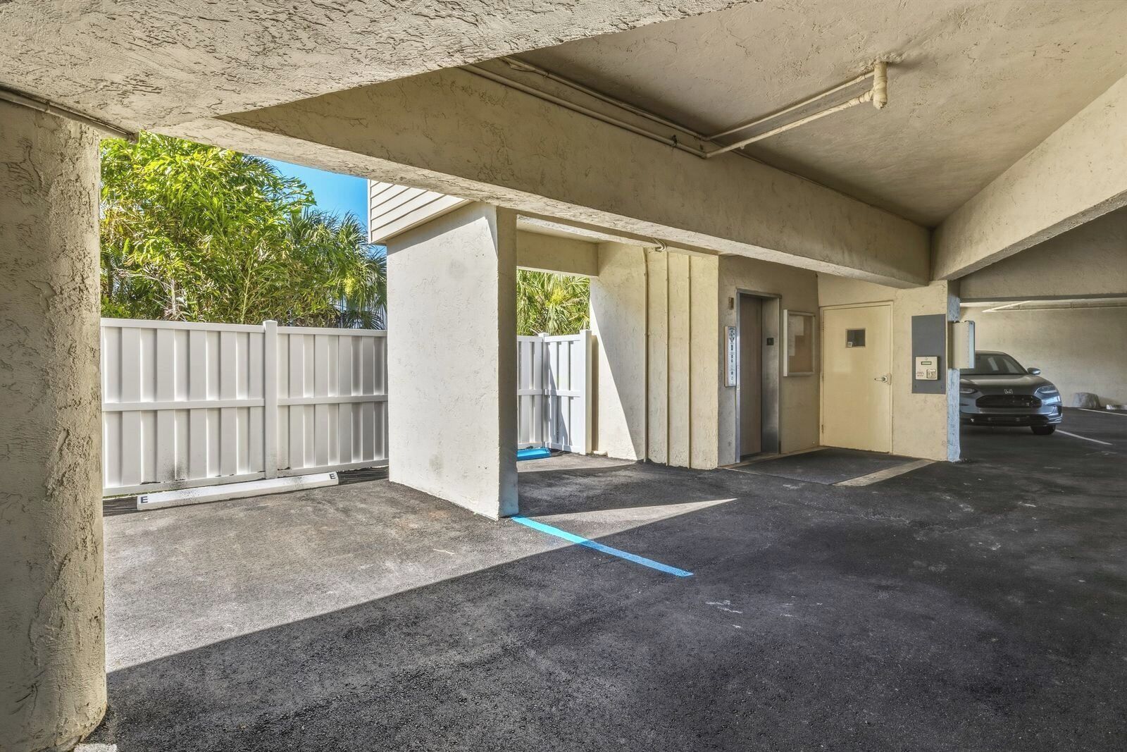 An indoor parking garage with concrete walls, a white fence outside, and a car parked in the distance.