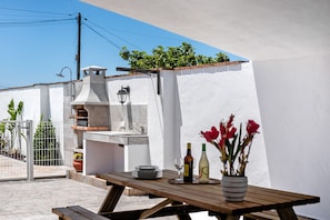 Un patio exterior con una parrilla, una mesa con platos y bebidas, y una planta en maceta.