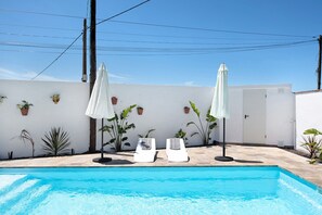 A swimming pool with two lounge chairs and umbrellas, surrounded by potted plants and a white wall.