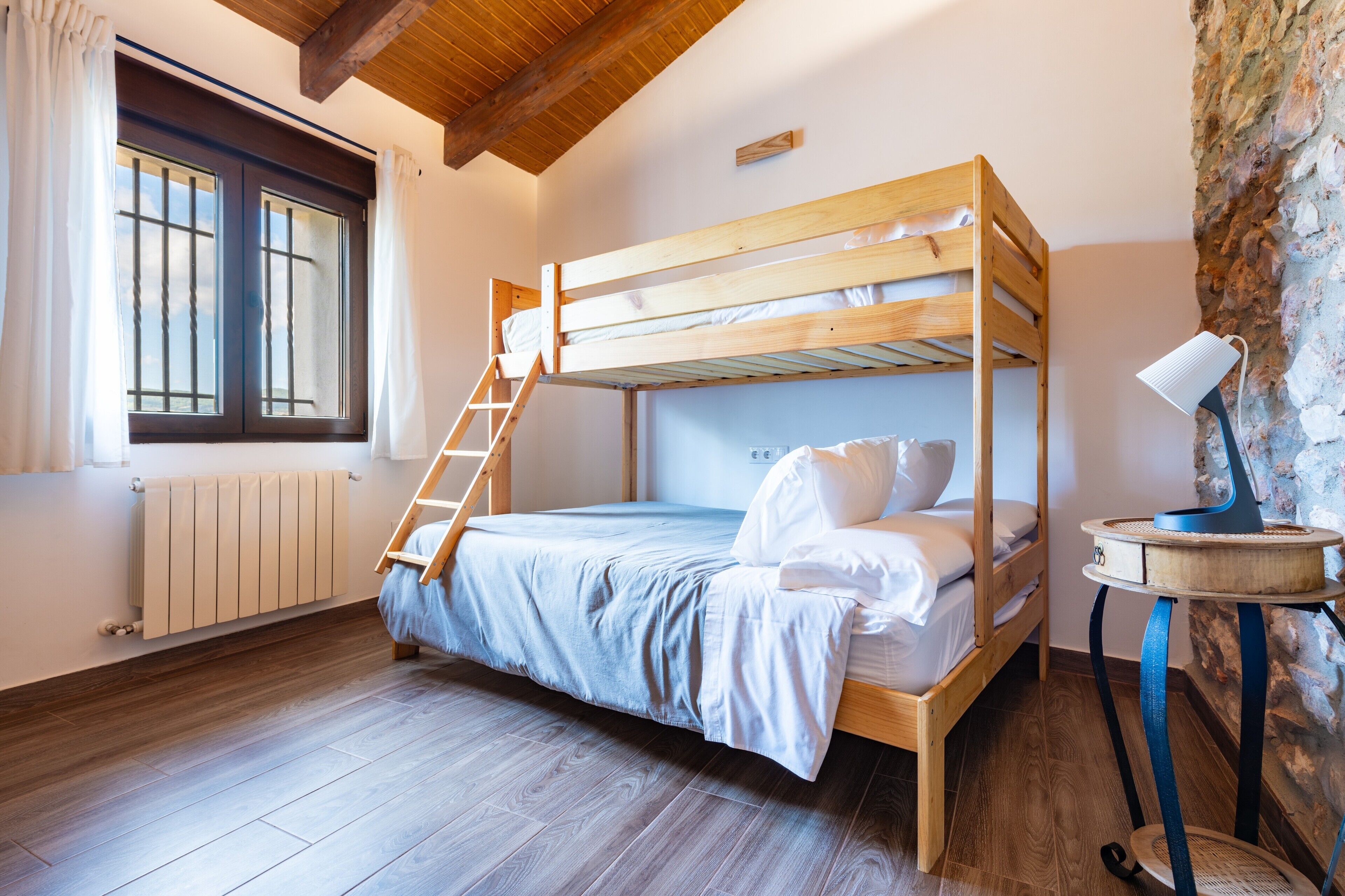 A bunk bed room with a wooden ladder, a small table with a lamp, and a window with curtains.