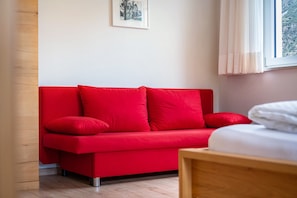 Un canapé rouge, un lit, un tableau encadré au mur et une fenêtre avec des rideaux.