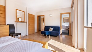 Moderni hotellihuone, jossa on sänky, sohva, tuoli, puinen työpöytä, peili ja näkymä viereiseen huoneeseen.