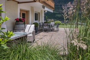 Een terras met tuinmeubilair, een grote plantenbak met bloemen en uitzicht op een bergachtig landschap.