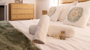 Una cama bien hecha con sábanas blancas y almohadas decorativas, un aparador de mimbre y un calentador fijado a la pared.