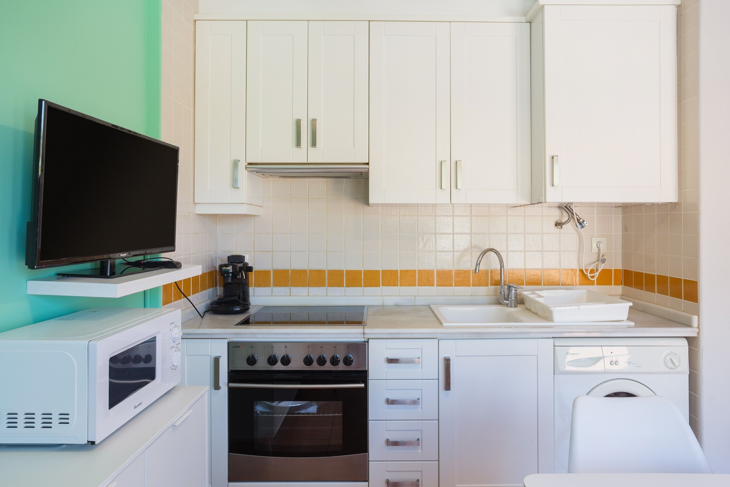 A compact kitchen with white cabinets, a microwave, an oven, a dishwasher, a sink, and a television.