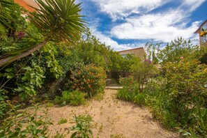 Um jardim com um caminho de terra, diversas plantas e um edifício ao fundo.