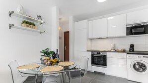Una cocina moderna con armarios blancos, horno empotrado, lavadora y comedor con mesa y sillas de vidrio.