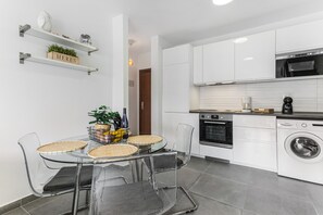 Une cuisine moderne avec des meubles blancs, un four encastré, un lave-linge et un coin repas comprenant une table et des chaises en verre.