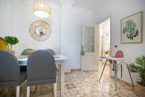 Uma área de refeições com uma mesa branca, cadeiras cinzas, um lustre pendurado, um espelho e uma porta que dá para outro cômodo.