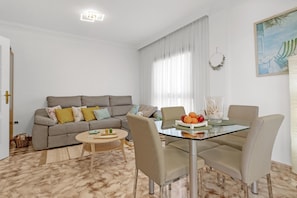 Una sala de estar con un sofá, una mesa de vidrio para comedor con sillas, una mesita redonda y un ventanal con cortinas.