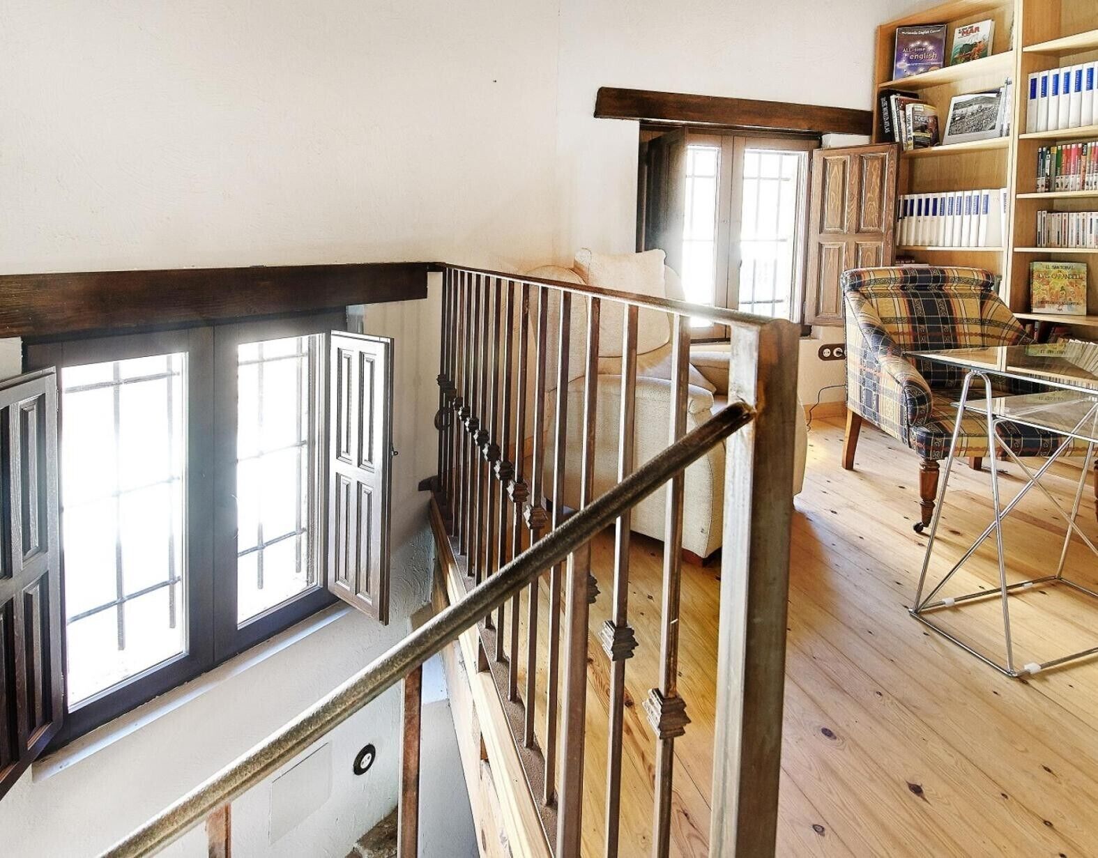 Una escalera con barandilla de metal, escalones de madera y vistas a una habitación con una estantería y un sillón a cuadros.