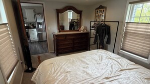 A bedroom with a large bed, a dresser, a mirror, a coat rack, and a view of the kitchen through an open door. - Maple Creek Retreat (Owatonna)