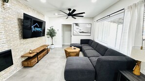 A modern living room with a sectional sofa, a flat-screen TV mounted on a brick wall, a ceiling fan, and a potted plant. - Beautiful 3 Bedroom 3 Bath Home w/ Pool Access (Fort Lauderdale)