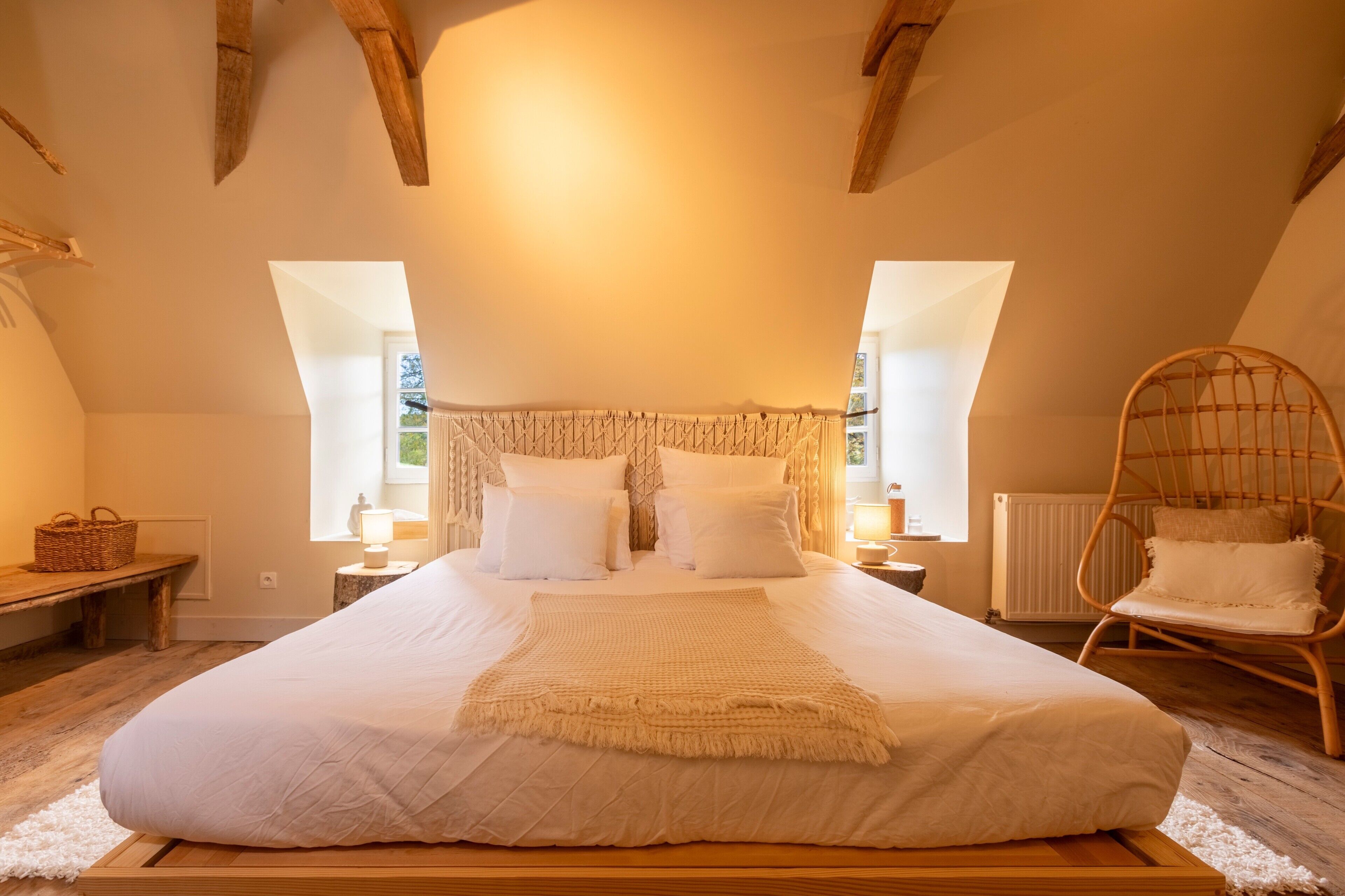 Une chambre avec un grand lit, des poutres en bois, un fauteuil en osier et une fenêtre donnant sur un beau paysage.