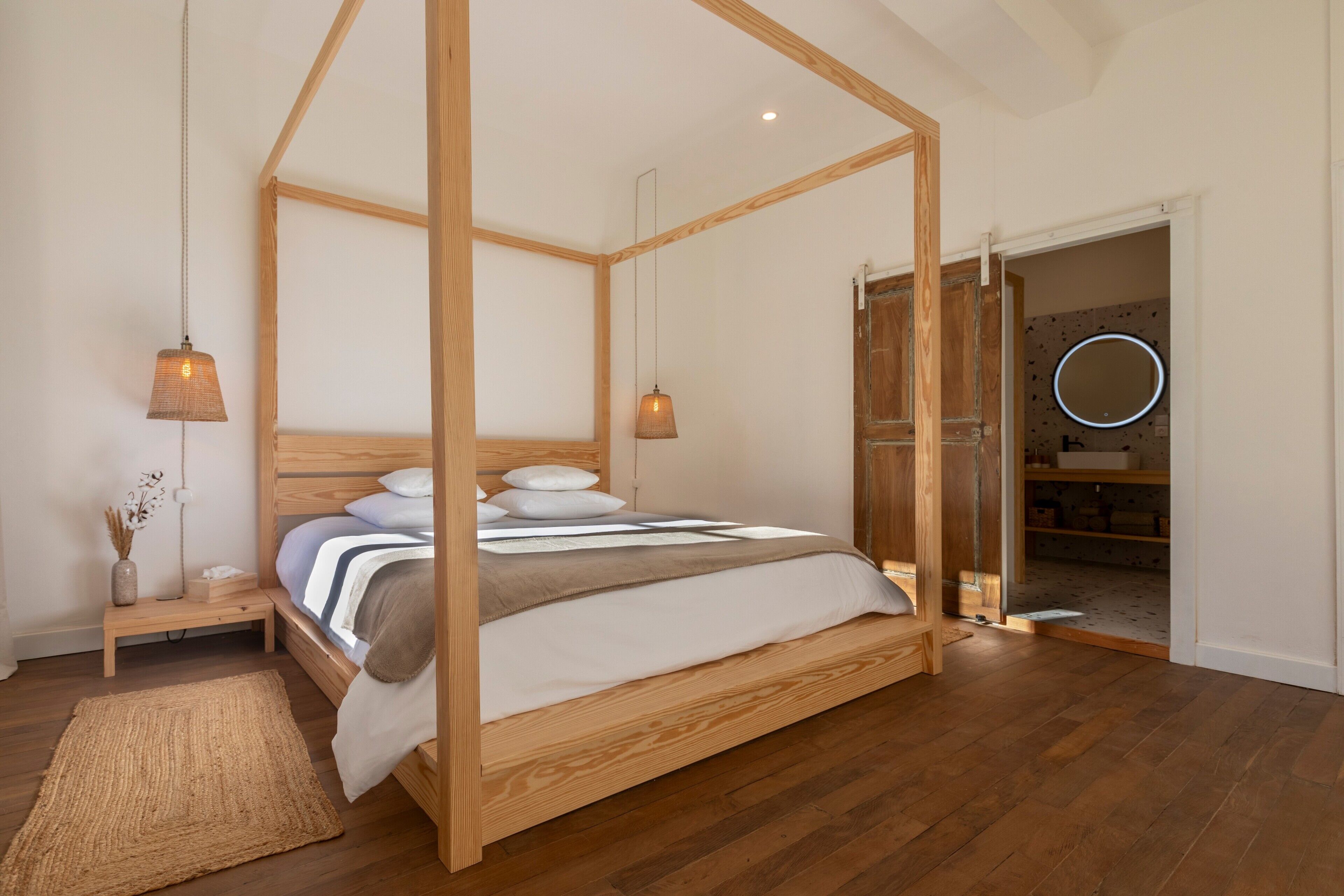 A four-poster bed with a wooden frame, a bedside table with a vase, and a mirror with a circular light.