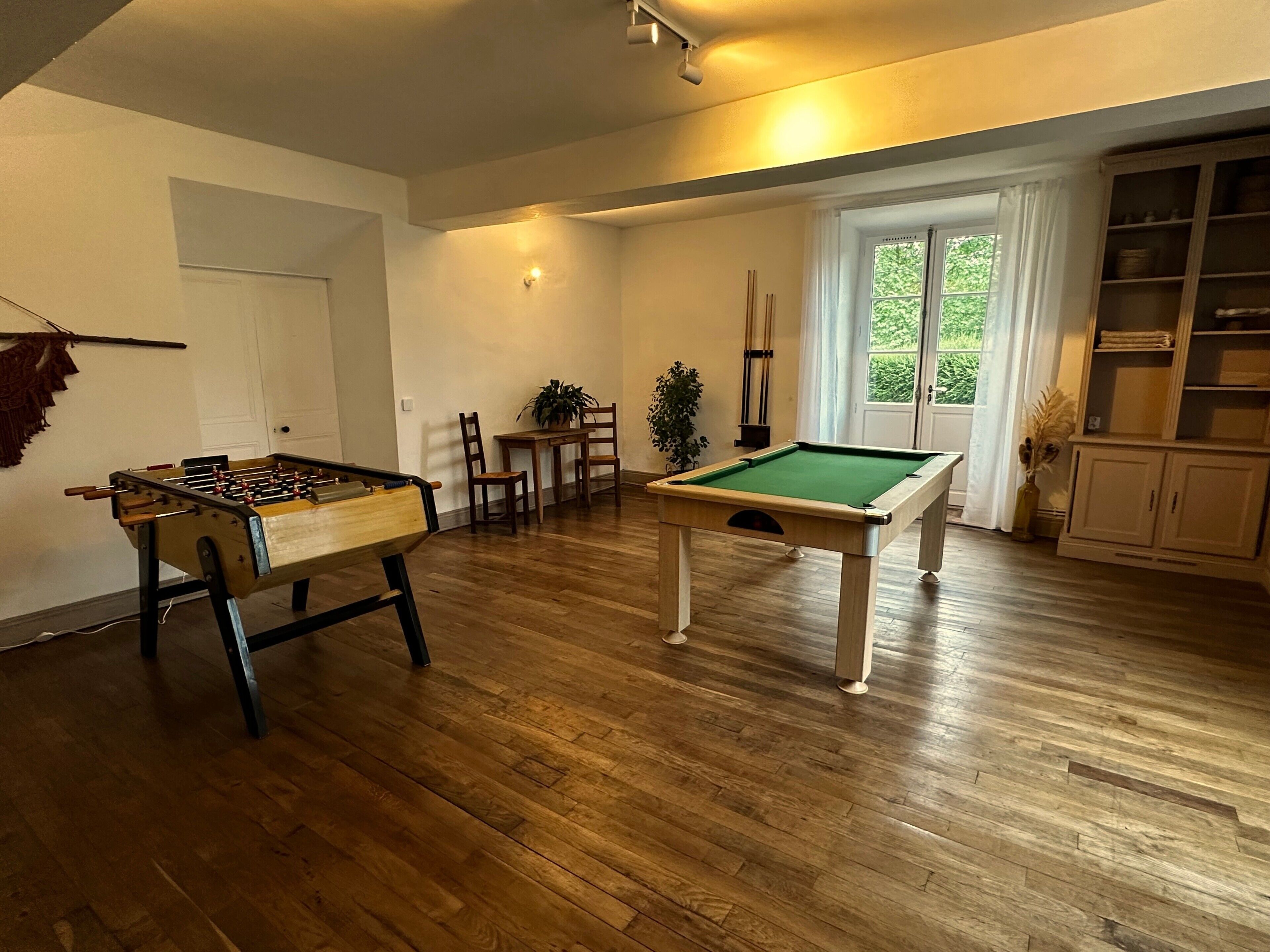 A room with a pool table, a foosball table, a dining area with chairs and a table, and a bookshelf.