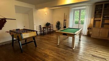 A room with a pool table, a foosball table, a dining area with chairs and a table, and a bookshelf.
