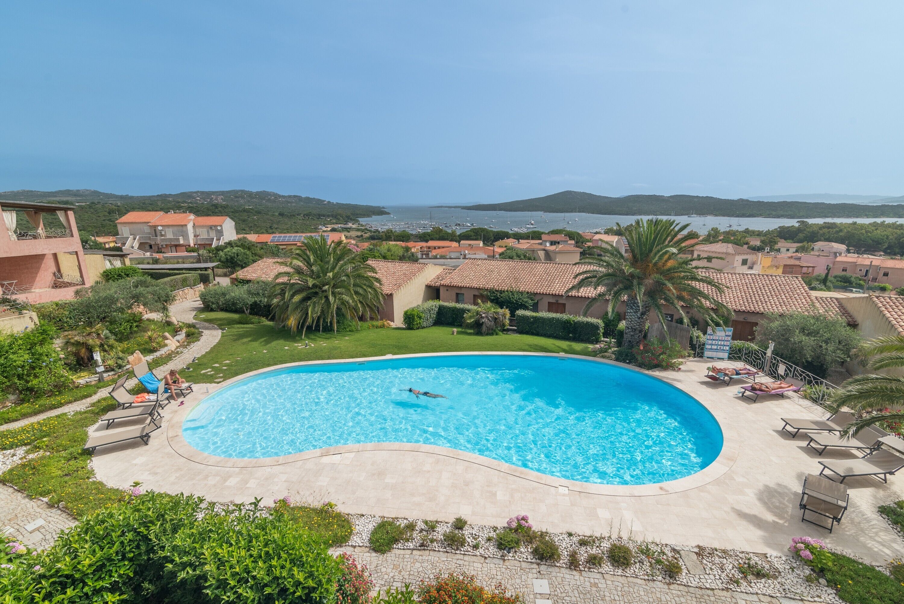 Une piscine à bordures courbes, entourée de chaises longues et de palmiers, offrant une vue sur une ville côtière et des collines en arrière-plan.