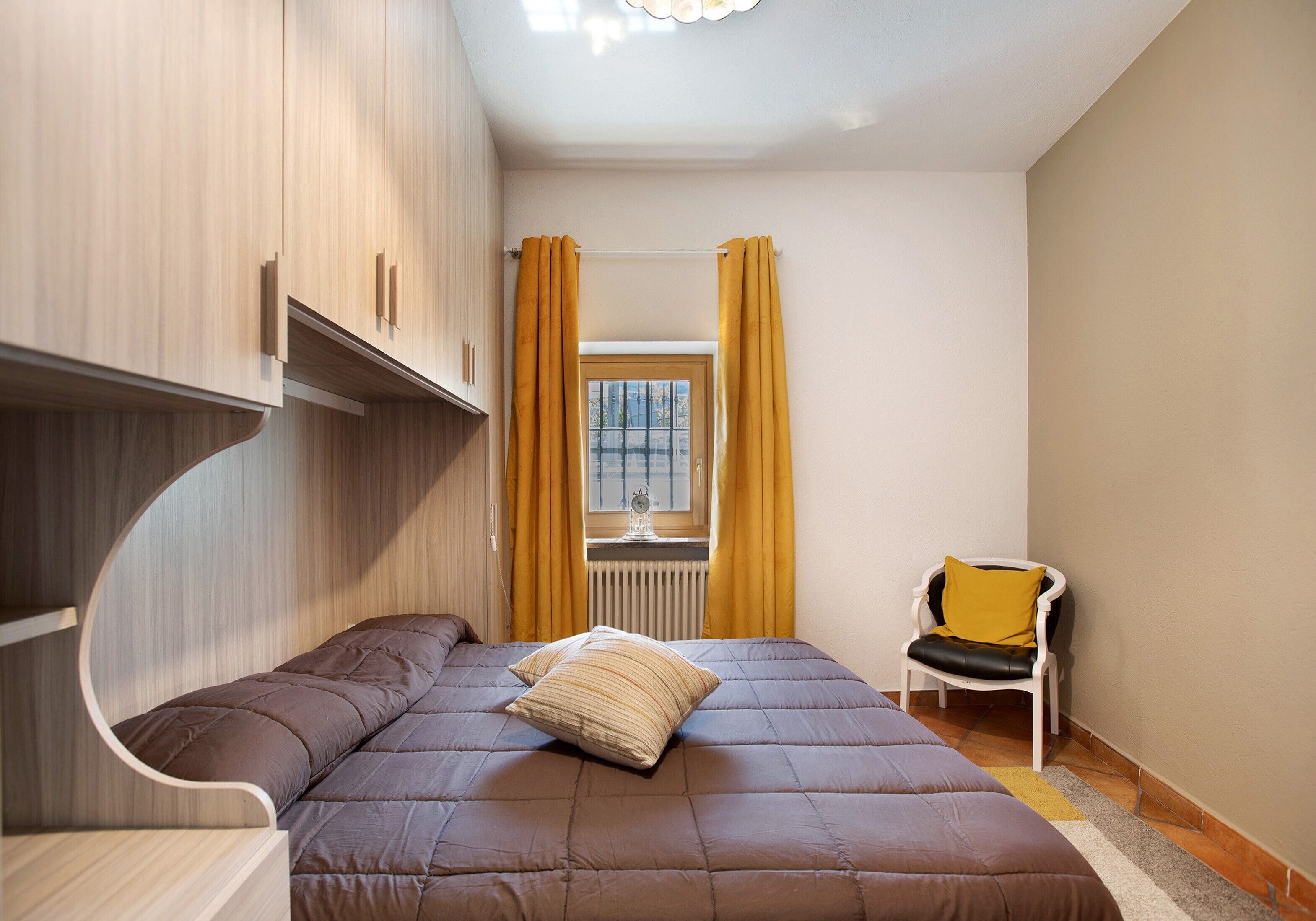 A bedroom with a bed, a chair, a window with curtains, and built-in wooden cabinets.