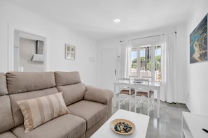Un salon avec un canapé beige, une table blanche sur laquelle se trouve un bol de collations, et une vue sur un balcon agrémenté de rideaux blancs.