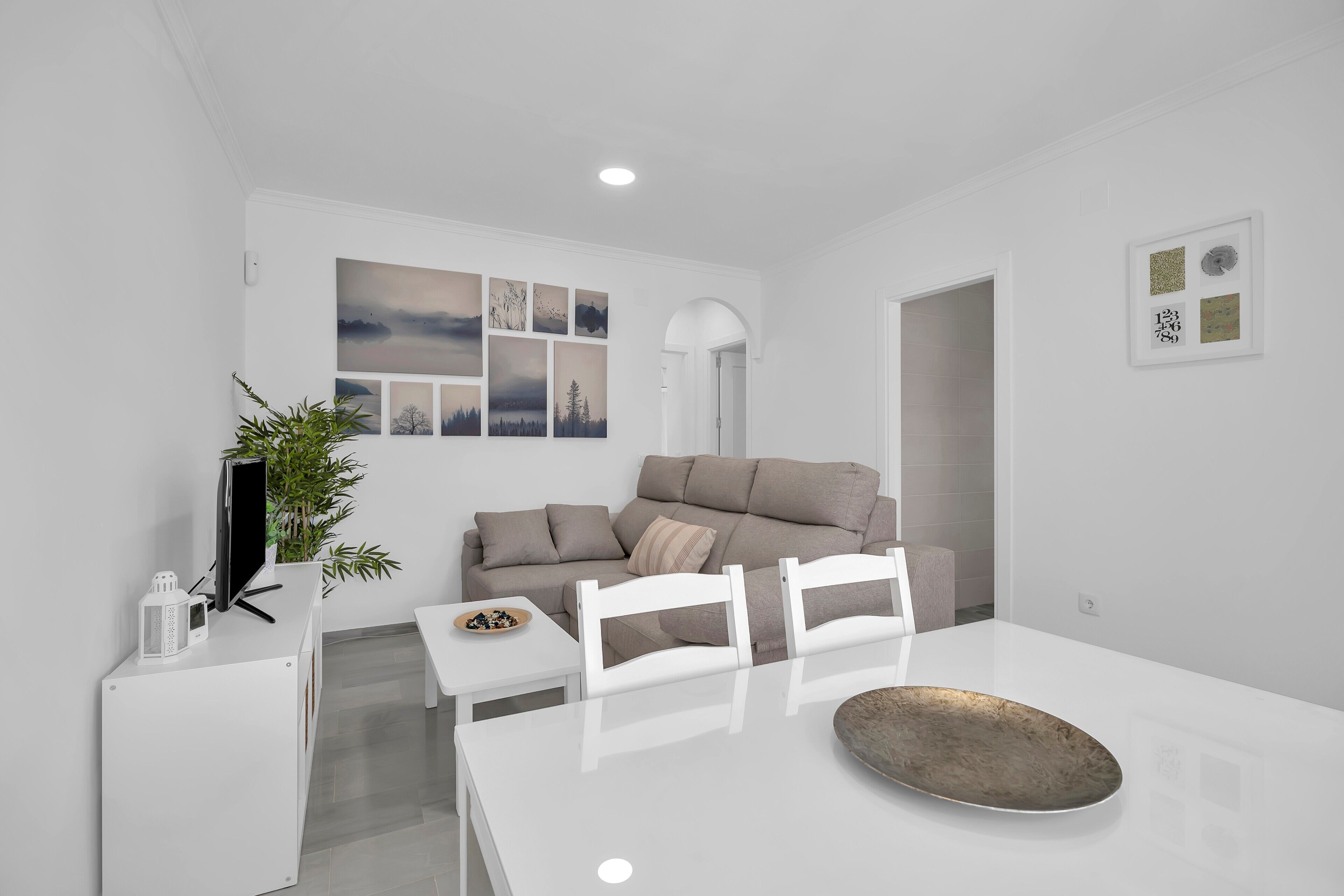 A modern living room with a white sofa, a dining table with chairs, a flat-screen TV, a potted plant, and artwork on the wall.