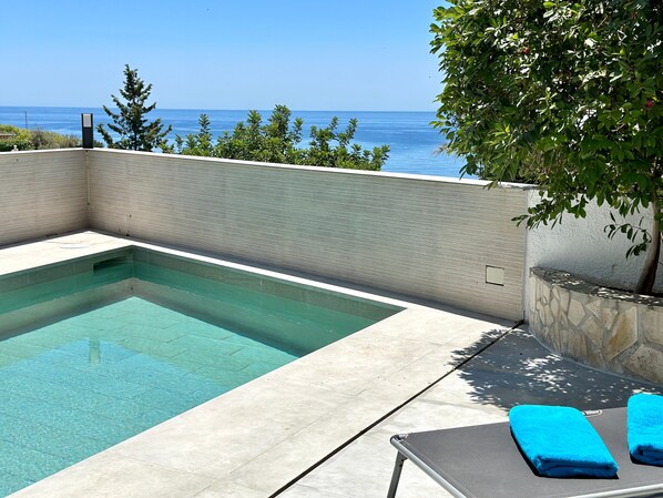 A swimming pool with a view of the ocean, a lounge chair with a blue towel, and a tree in the background.