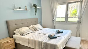 Ein Schlafzimmer mit einem Bett, Nachttischen, einer Bank, einem Fenster mit Vorhängen und an der Wand befestigten Regalen mit Pflanzen.