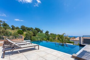 A swimming pool with a wooden deck and lounge chairs, surrounded by greenery and a view of the sea.