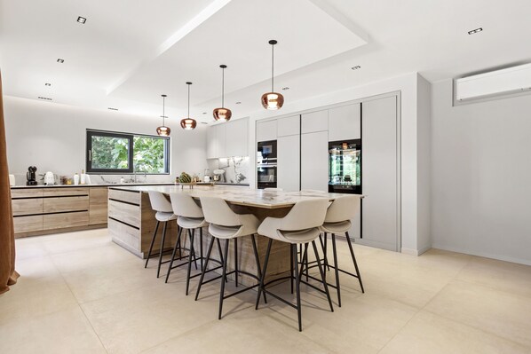 A modern kitchen with a central island, bar stools, pendant lighting, and a large window.