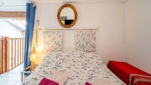 A bedroom with a bed, a mirror, a red chair, a lamp, and a window with a view of a balcony.