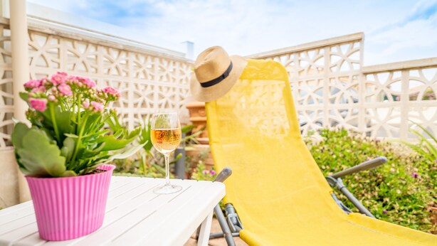 A white table with a pink pot of flowers, a glass of wine, and a yellow chair.