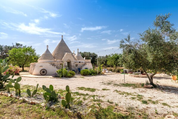 A traditional stone building with a conical roof, surrounded by cacti and olive trees.