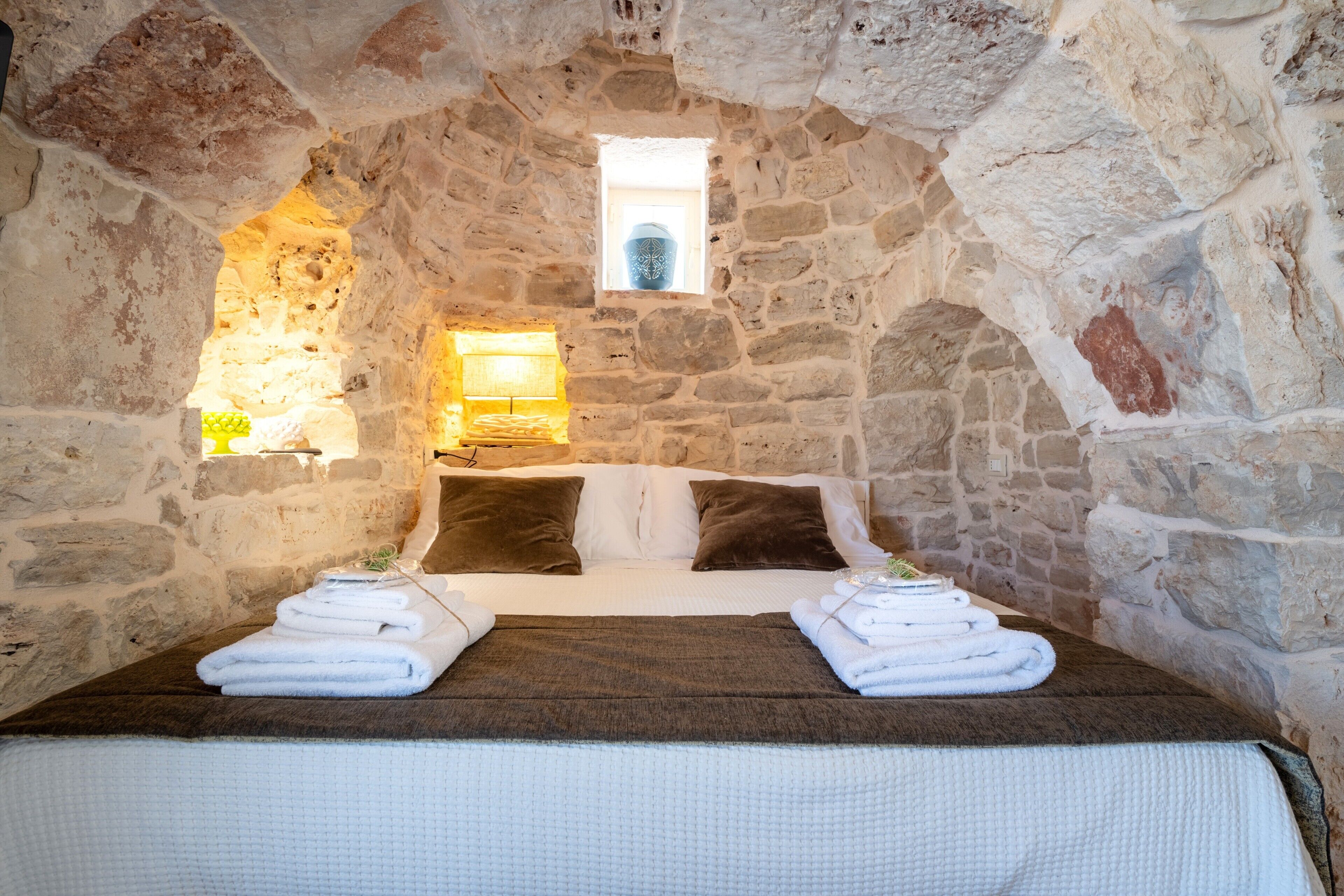 A stone-walled room with a bed, two pillows, a window, and a nightstand with a lamp.