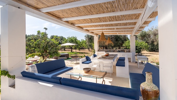 Un patio al aire libre con mobiliario blanco, cojines azules, una mesa de vidrio y un jarrón grande.