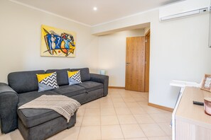 Una sala de estar con un sofá gris, un cuadro abstracto de colores, una puerta de madera y un equipo de aire acondicionado blanco.