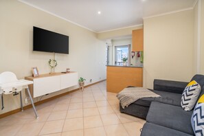 Una sala de estar con televisor de pantalla plana, un sofá gris, una trona blanca y una zona de cocina al fondo.