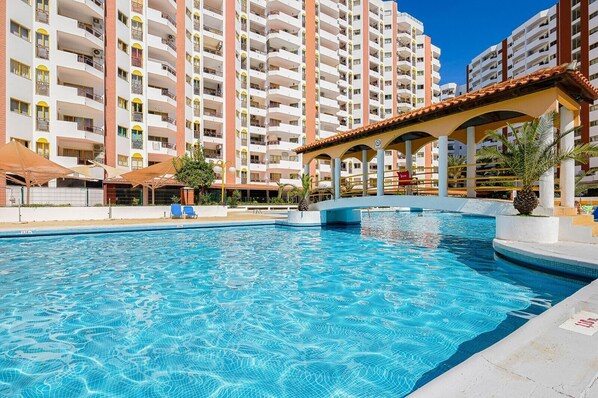 Une piscine à l’eau d’un bleu cristallin, entourée d’un bâtiment blanc doté de balcons et d’un espace abrité avec des parasols.
