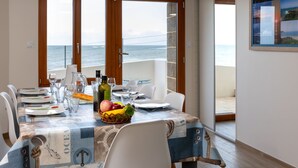 A dining area with a view of the sea, a table set with plates, glasses, and a fruit basket.