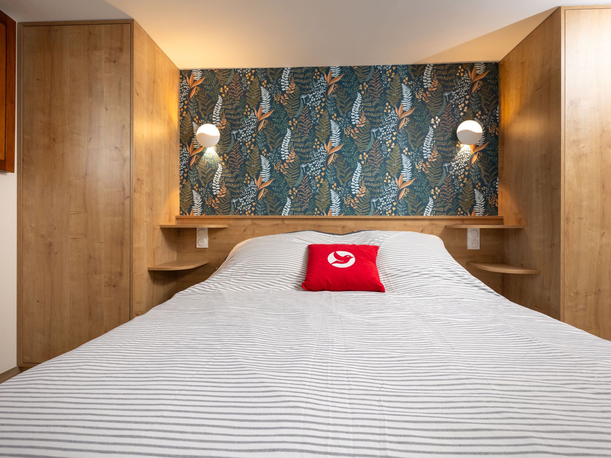 A hotel room with a bed, a red pillow, wooden walls, and a floral wallpaper.