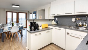 A modern kitchen with white cabinets, a grey countertop, and a view of the sea through large windows.