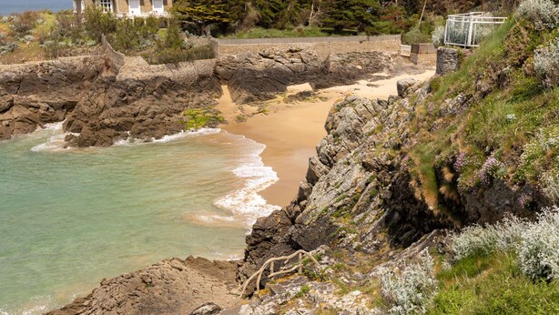 A coastal landscape with rocky cliffs, a sandy beach, and a clear view of the sea.