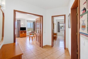 A room with a wooden door, a wooden bench, a television, and a view through an open door to another room with a window.