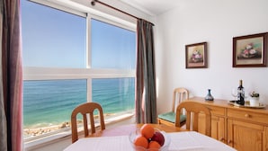 A room with a large window overlooking the sea, a dining table with a fruit bowl, and a wooden cabinet with a vase and wine glasses.