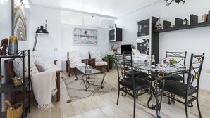 Una sala de estar con un sofá, una mesa de centro, una mesa de comedor con sillas, una araña y un televisor.