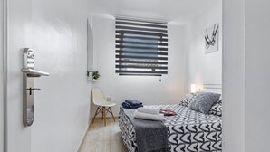 Un dormitorio con cama, una silla, un ventanal con persianas, una puerta y una obra de arte colgada en la pared.
